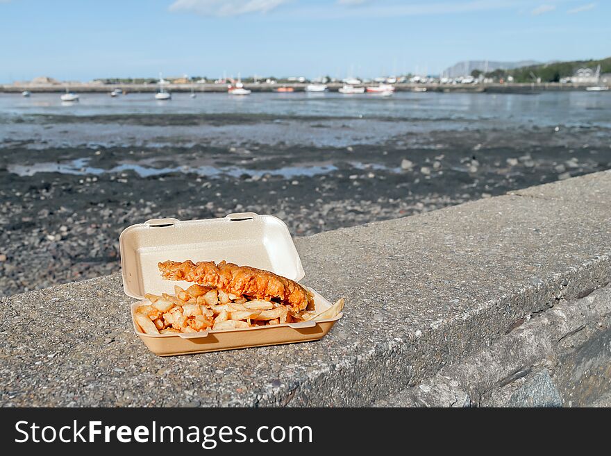 A tray of fish and chips on a harbour wall showing out of focus boats in the background