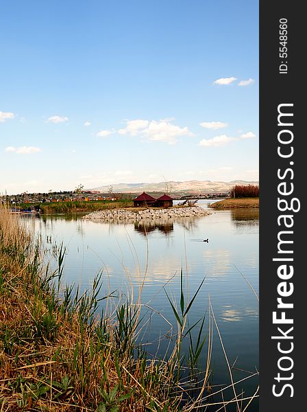 A serene view of a pond in Mogan Park near Ankara, the capital of Turkey, displaying its blue surface framed with reeds and the bright blue sky with clouds. A serene view of a pond in Mogan Park near Ankara, the capital of Turkey, displaying its blue surface framed with reeds and the bright blue sky with clouds