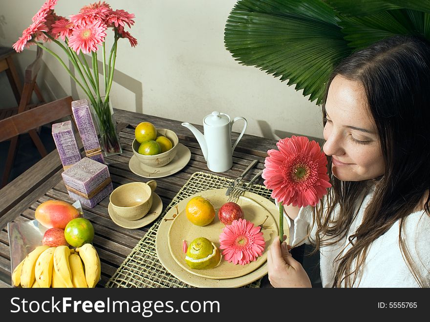 An attractive young girl outside, smelling a pink flower, sitting at a table, while fruits and plants surround her. - horizontally framed. An attractive young girl outside, smelling a pink flower, sitting at a table, while fruits and plants surround her. - horizontally framed