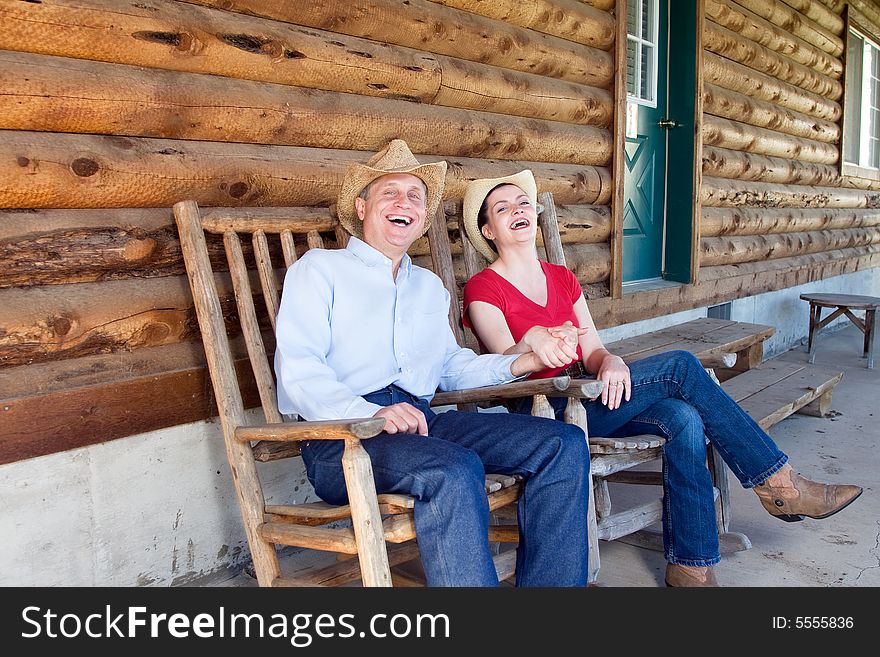 A Cowgirl and cowboy laughing and holding hands as they sit together on a porch.  Horizontally framed photograph. A Cowgirl and cowboy laughing and holding hands as they sit together on a porch.  Horizontally framed photograph