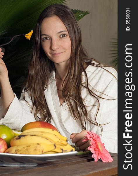 A young girl, sitting at a table full of fruits in a white bathrobe eating a mango. - vertically framed. A young girl, sitting at a table full of fruits in a white bathrobe eating a mango. - vertically framed