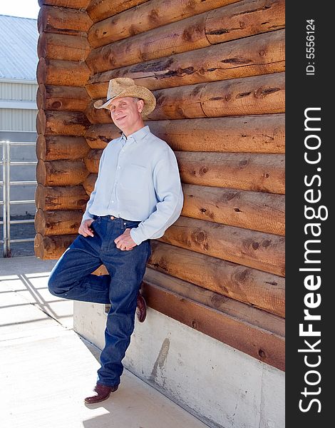 Man in cowboy hat leaning against a log cabin with his leg propped up. Verictally framed photograph. Man in cowboy hat leaning against a log cabin with his leg propped up. Verictally framed photograph