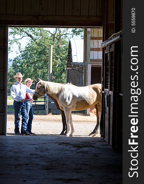 A Cowgirl and Cowboy smiling as they pet a brown horse outside a barn. Hoizontally framed photograph. A Cowgirl and Cowboy smiling as they pet a brown horse outside a barn. Hoizontally framed photograph