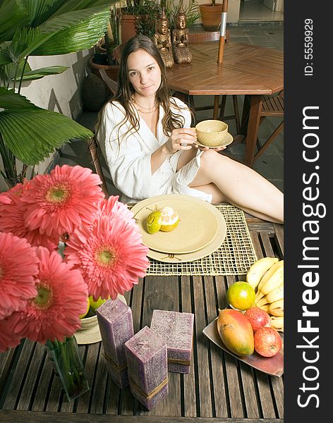 A young girl sitting at a table with plants, fruits, and flowers on it. She is holding a cup while looking at the camera. - vertically framed. A young girl sitting at a table with plants, fruits, and flowers on it. She is holding a cup while looking at the camera. - vertically framed