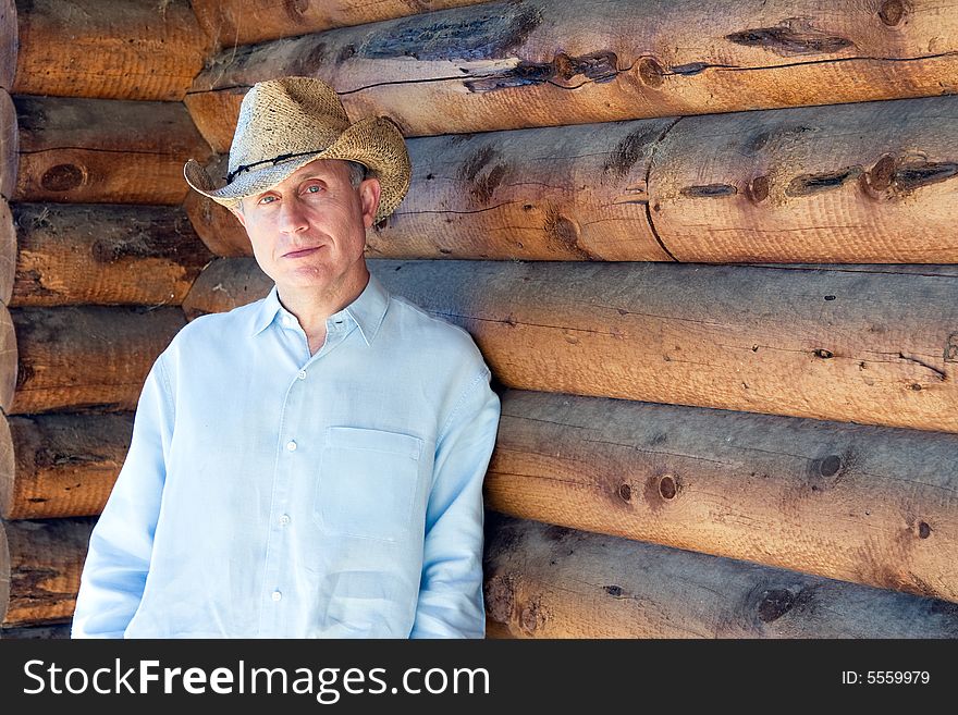 Man in cowboy hat leaning against a log cabin. Horizontally framed photograph. Man in cowboy hat leaning against a log cabin. Horizontally framed photograph