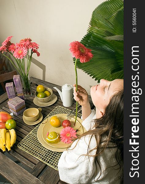 An attractive young girl, smelling a pink flower, sitting at a table, while fruits and plants surround her. - vertically framed. An attractive young girl, smelling a pink flower, sitting at a table, while fruits and plants surround her. - vertically framed