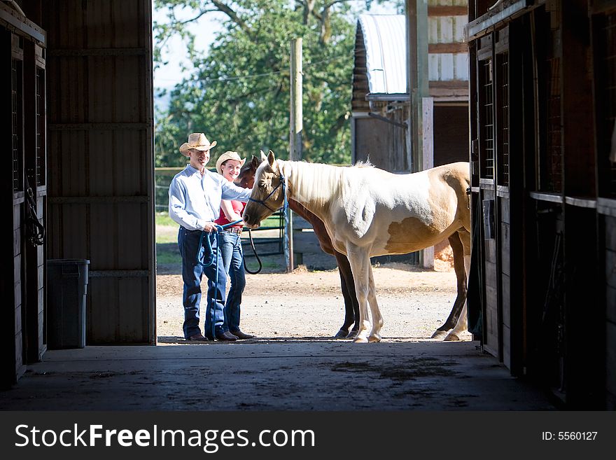 A Cowgirl and Cowboy smiling as they pet a brown horse outside a barn. Hoizontally framed photograph. A Cowgirl and Cowboy smiling as they pet a brown horse outside a barn. Hoizontally framed photograph