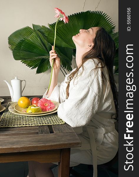 An attractive young girl outside, staring at a pink flower, sitting at a table, while fruits and plants surround her. - vertically framed. An attractive young girl outside, staring at a pink flower, sitting at a table, while fruits and plants surround her. - vertically framed