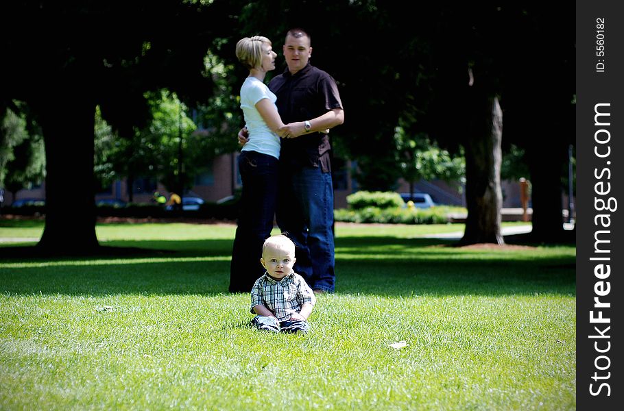 The parents of the baby are standing behing the baby, hugging. The baby is seated in the grass, laughing. - horizontally framed. The parents of the baby are standing behing the baby, hugging. The baby is seated in the grass, laughing. - horizontally framed