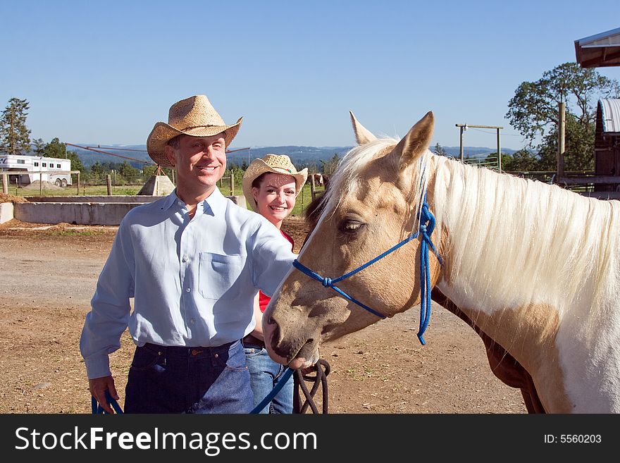 A Cowgirl and Cowboy smiling as they pet a blonde horse. Hoizontally framed photograph. A Cowgirl and Cowboy smiling as they pet a blonde horse. Hoizontally framed photograph