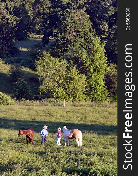 Two people holding their horses, standing in a meadow of grass, with trees. - vertically framed. Two people holding their horses, standing in a meadow of grass, with trees. - vertically framed