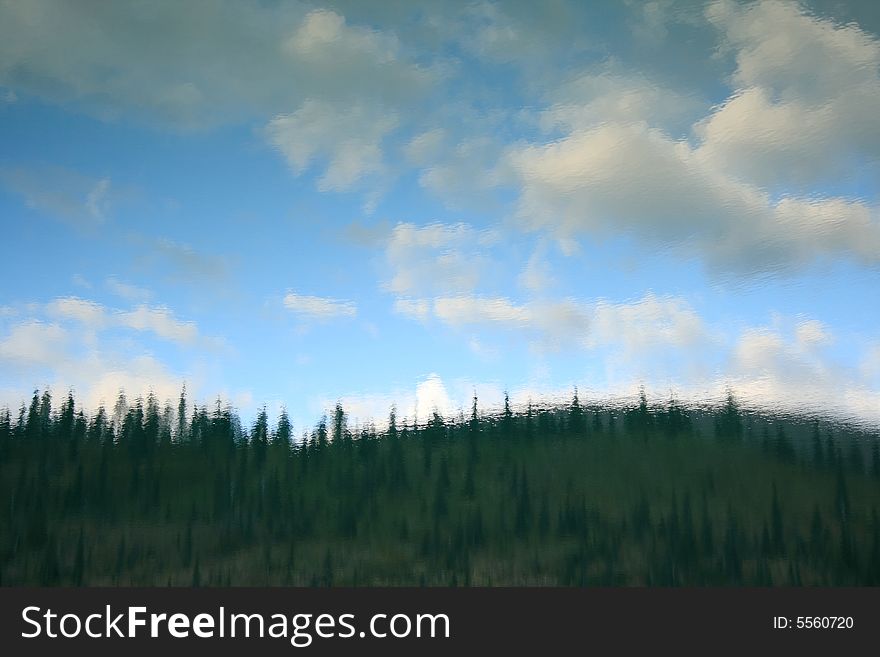 Reflection of a forested shoreline and blue sky with clouds reflecting from a calm lake to create a painterly effect. Reflection of a forested shoreline and blue sky with clouds reflecting from a calm lake to create a painterly effect