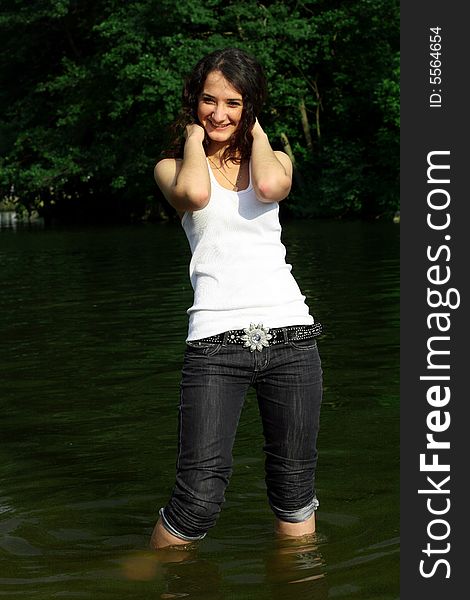 The girl in dark blue jeans and a white vest costs in water on a background of green trees. The girl in dark blue jeans and a white vest costs in water on a background of green trees