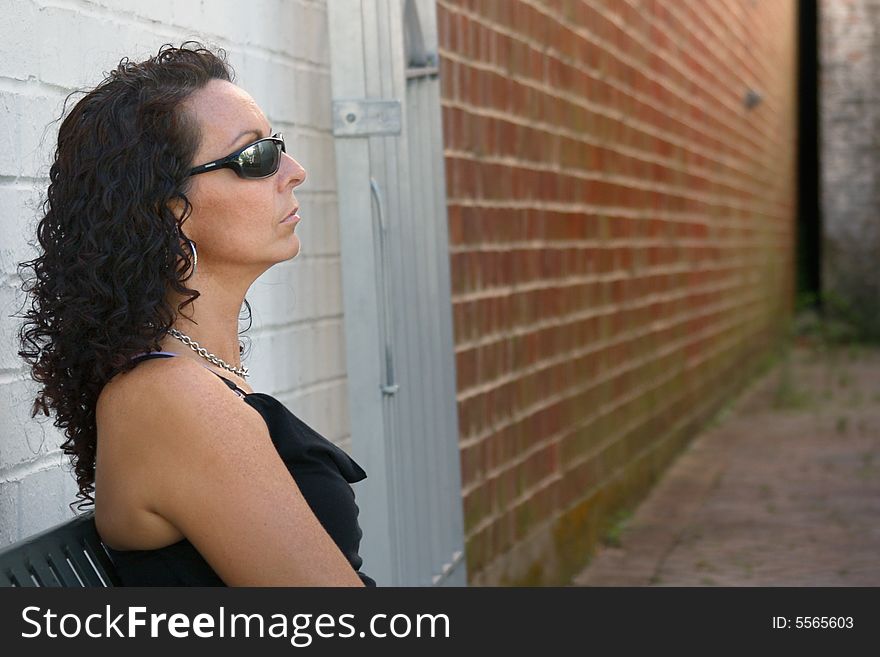 A pretty woman sitting outside of a long empty alley near a red brick wall.