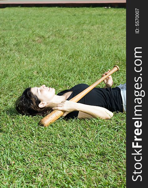 A woman is laying on the grass at the park. She is holding a baseball bat across her chest, and appears to be sleeping or resting. Vertically framed shot. A woman is laying on the grass at the park. She is holding a baseball bat across her chest, and appears to be sleeping or resting. Vertically framed shot.