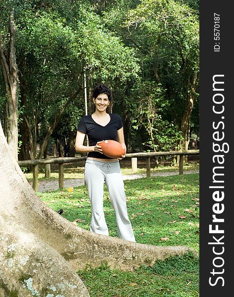 A young, attractive woman is standing next to a tree at the park.  She is smiling and holding a football.Vertically framed photo. A young, attractive woman is standing next to a tree at the park.  She is smiling and holding a football.Vertically framed photo.