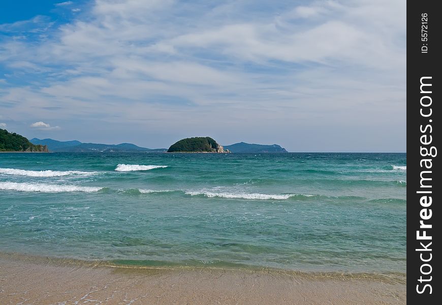 A small island on Japanese sea. On foreground is surf, on background is sky with white clouds. Russian Far East, state narure reserve Lazovsky, Beltsova island. A small island on Japanese sea. On foreground is surf, on background is sky with white clouds. Russian Far East, state narure reserve Lazovsky, Beltsova island.