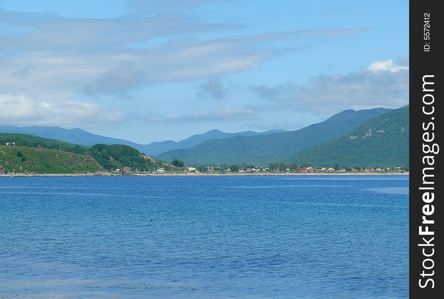 A landscape at sea. A blue seawater on foreground. A sky with white clouds, green mountaines and village at bank. Russian Far East, Primorye, Sokolovka village. A landscape at sea. A blue seawater on foreground. A sky with white clouds, green mountaines and village at bank. Russian Far East, Primorye, Sokolovka village.