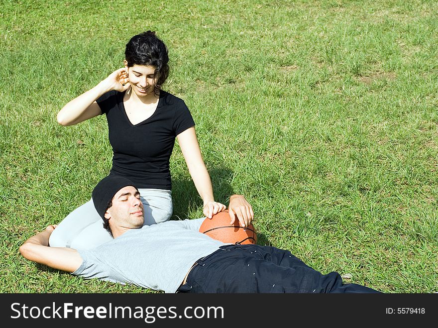 Young, attractive couple sitting together on a grassy field.  The man is laying across the woman's lap and holding up a basketball.  Horizontally framed shot. Young, attractive couple sitting together on a grassy field.  The man is laying across the woman's lap and holding up a basketball.  Horizontally framed shot.