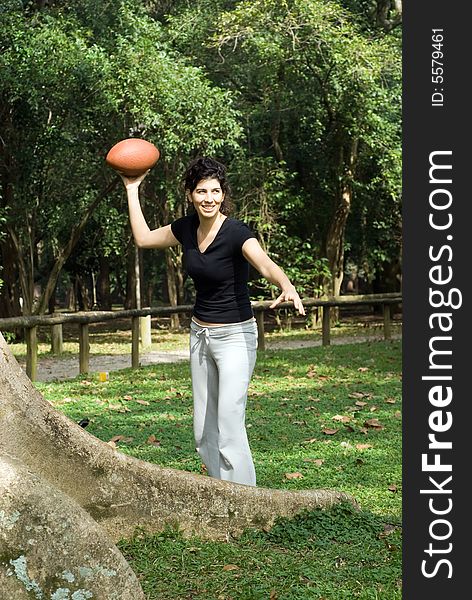 A young, attractive woman is standing next to a tree at the park.  She is smiling and about to throw a football.Vertically framed photo. A young, attractive woman is standing next to a tree at the park.  She is smiling and about to throw a football.Vertically framed photo.