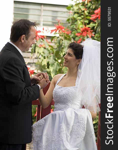 A newly married couple smile at each other while clasping hands, red flowers urban building with red flowers  in background. - vertically framed. A newly married couple smile at each other while clasping hands, red flowers urban building with red flowers  in background. - vertically framed