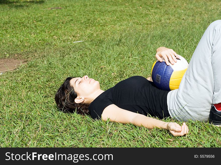 A woman is laying on the grass at the park. She is holding a volleyball on her stomach, and appears to be sleeping or resting. Horizontally framed shot. A woman is laying on the grass at the park. She is holding a volleyball on her stomach, and appears to be sleeping or resting. Horizontally framed shot.