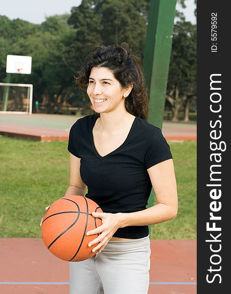 Young, attractive, happy woman is standing on an outdoor basketball court.  She is holding a basketball and looking in the other direction.  Vertically framed shot. Young, attractive, happy woman is standing on an outdoor basketball court.  She is holding a basketball and looking in the other direction.  Vertically framed shot.