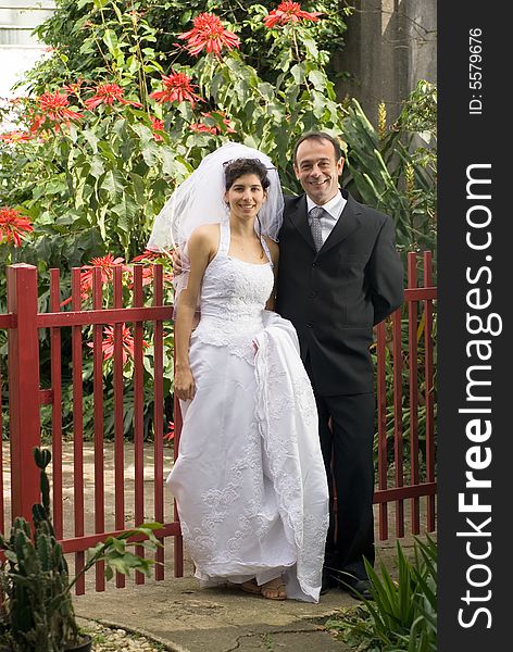 Married couple pose in front of a red fence, with red flowers in background. - vertically framed. Married couple pose in front of a red fence, with red flowers in background. - vertically framed