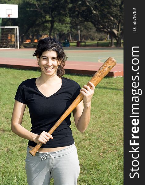 A smiling woman in a park is holding a baseball bat.  Vertically framed shot. A smiling woman in a park is holding a baseball bat.  Vertically framed shot.