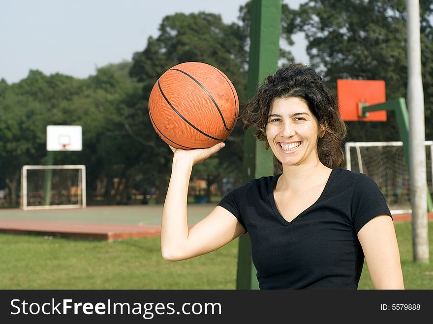 A woman smiling for the camera with a basketball court in the background, holding a basketball in one hand. - horizontally framed. A woman smiling for the camera with a basketball court in the background, holding a basketball in one hand. - horizontally framed