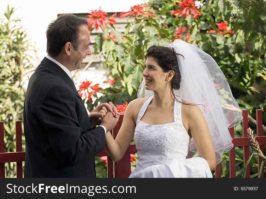 A newly married couple smile at each other while holding hands, red flowers predominant in background. - horizontally framed. A newly married couple smile at each other while holding hands, red flowers predominant in background. - horizontally framed