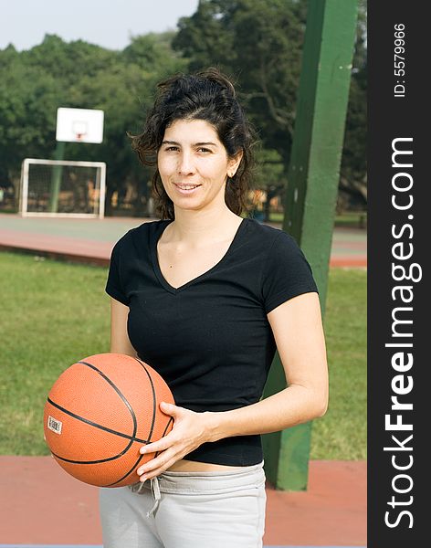 Young, attractive, happy woman is standing on an outdoor basketball court.  She is holding a basketball and looking at the camera.  Vertically framed shot. Young, attractive, happy woman is standing on an outdoor basketball court.  She is holding a basketball and looking at the camera.  Vertically framed shot.