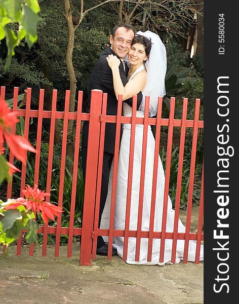 A couple, newly married, smile for the camera while hugging behind a red gate. - vertically framed. A couple, newly married, smile for the camera while hugging behind a red gate. - vertically framed