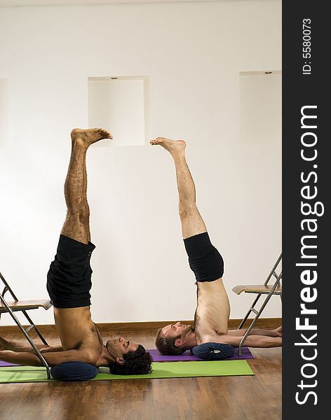 Two men performing a candlestick while on mats, against chairs. - vertically framed. Two men performing a candlestick while on mats, against chairs. - vertically framed