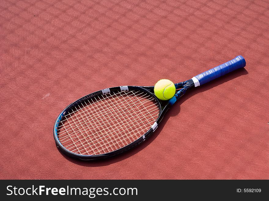 A set of tennis racket and green ball placed on the red tennis court with shadow. A set of tennis racket and green ball placed on the red tennis court with shadow.