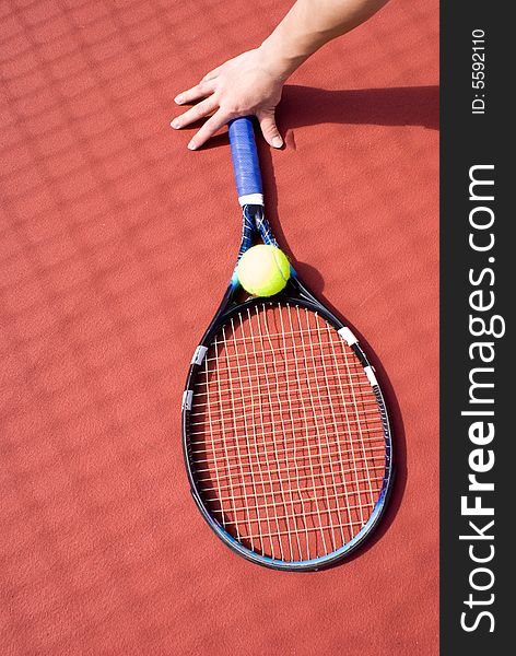 A hand holding the tennis racket with a green tennis ball on it resting at the gournd of the tennis court. A hand holding the tennis racket with a green tennis ball on it resting at the gournd of the tennis court.