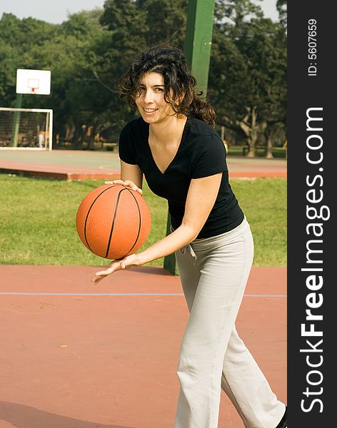 A woman is playing basketball on a park basketball court. She is smiling and looking at the camera. She looks like she is about to dribble the basketball. Vertically framed photo. A woman is playing basketball on a park basketball court. She is smiling and looking at the camera. She looks like she is about to dribble the basketball. Vertically framed photo.