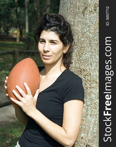 Young, attractive woman is leaning against a tree.  She is holding a football.   Vertically framed shot. Young, attractive woman is leaning against a tree.  She is holding a football.   Vertically framed shot