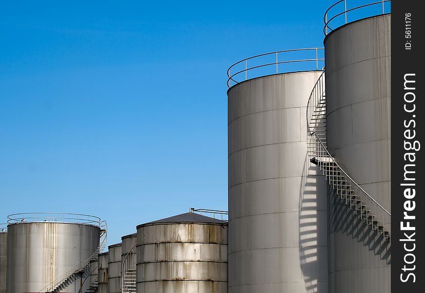 Row of cylindrical industrial storage tanks of various heights and sizes with spiral staircases on their sides leading to access on their roofs. Row of cylindrical industrial storage tanks of various heights and sizes with spiral staircases on their sides leading to access on their roofs