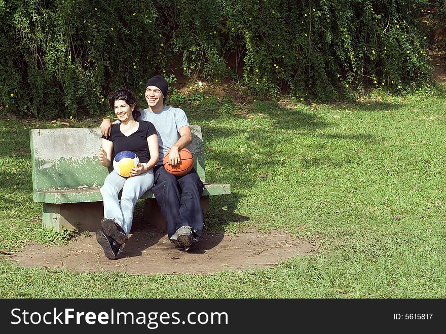 A couple is sitting on a park bench. They are both smiling and laughing and looking away from the camera and each other. The man is holding a basketball and the woman is holding a volleyball. Horizontally framed photo. A couple is sitting on a park bench. They are both smiling and laughing and looking away from the camera and each other. The man is holding a basketball and the woman is holding a volleyball. Horizontally framed photo.