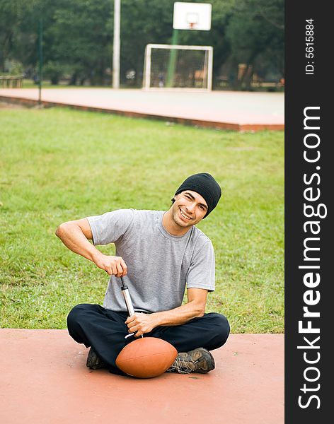 A man is sitting in a park. He is smiling, looking at the camera and pumping air into a football. Vertically framed photo. A man is sitting in a park. He is smiling, looking at the camera and pumping air into a football. Vertically framed photo.