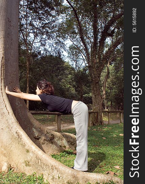 A young, attractive woman is standing next to a tree. She is looking at the tree, leaning against it and stretching. Vertically framed photo. A young, attractive woman is standing next to a tree. She is looking at the tree, leaning against it and stretching. Vertically framed photo.