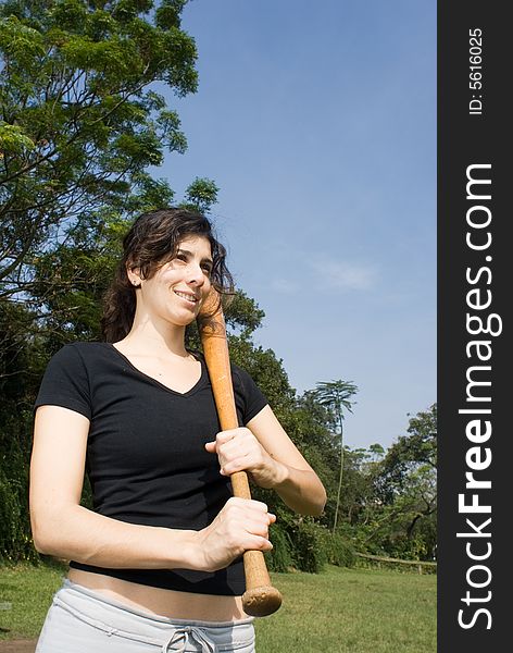 A young, attractive woman is standing in a park.  She is looking away from the camera and smiling.  She is posing with a baseball bat.  Vertically framed photo. A young, attractive woman is standing in a park.  She is looking away from the camera and smiling.  She is posing with a baseball bat.  Vertically framed photo.