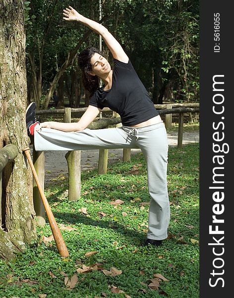 A smiling woman is standing next to a tree.  She is looking away from the camera and stretching.  There is a baseball bat leaning against the tree.  Vertically framed photo. A smiling woman is standing next to a tree.  She is looking away from the camera and stretching.  There is a baseball bat leaning against the tree.  Vertically framed photo.