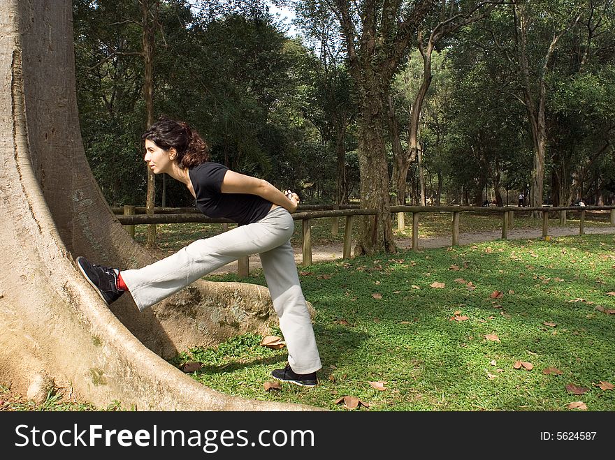 A young, attractive woman is standing next to a tree. She is looking at the tree, leaning against it and stretching. Horizontally framed photo. A young, attractive woman is standing next to a tree. She is looking at the tree, leaning against it and stretching. Horizontally framed photo.