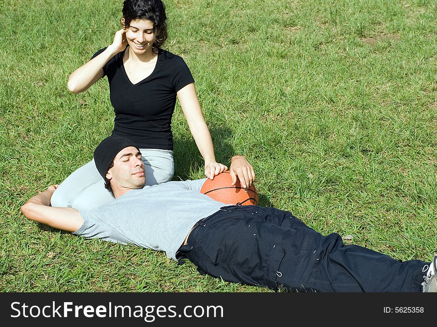 A couple is sitting on the grass in a park.  The woman is smiling and looking at the man and the man is resting on the womans lap.  The man is holding a basketball.  Horizontally framed photo. A couple is sitting on the grass in a park.  The woman is smiling and looking at the man and the man is resting on the womans lap.  The man is holding a basketball.  Horizontally framed photo.