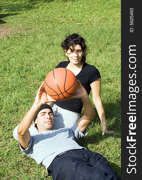 A couple is sitting on the grass in a park.  The woman is smiling and looking away from the camera and the man is resting on the womans lap.  The man is holding a basketball above his chest.  Vertically framed photo. A couple is sitting on the grass in a park.  The woman is smiling and looking away from the camera and the man is resting on the womans lap.  The man is holding a basketball above his chest.  Vertically framed photo.