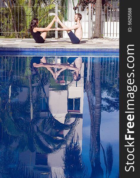 A couple, with their feet touching and hands linked, stretch with each other next to the pool - vertically framed. A couple, with their feet touching and hands linked, stretch with each other next to the pool - vertically framed
