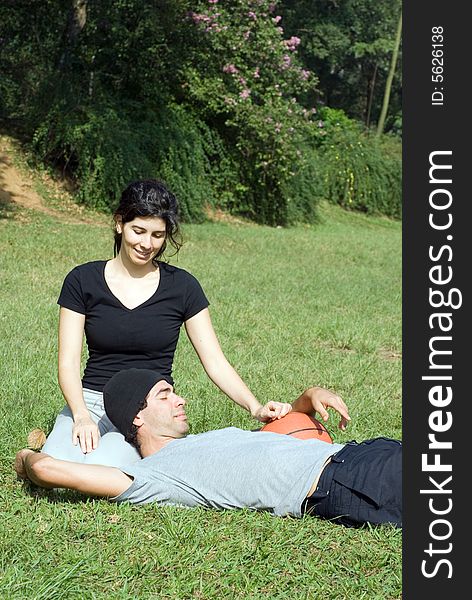 A couple is sitting on the grass in a park. The woman is smiling and looking at the man and the man is resting on the womans lap. The man is holding a basketball. Vertically framed photo. A couple is sitting on the grass in a park. The woman is smiling and looking at the man and the man is resting on the womans lap. The man is holding a basketball. Vertically framed photo.