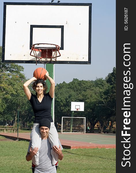 A couple is playing on a basketball court.  The woman is sitting on the man's shoulders and holding a basketball.  They are smiling and looking at the camera.  Vertically framed photo. A couple is playing on a basketball court.  The woman is sitting on the man's shoulders and holding a basketball.  They are smiling and looking at the camera.  Vertically framed photo.